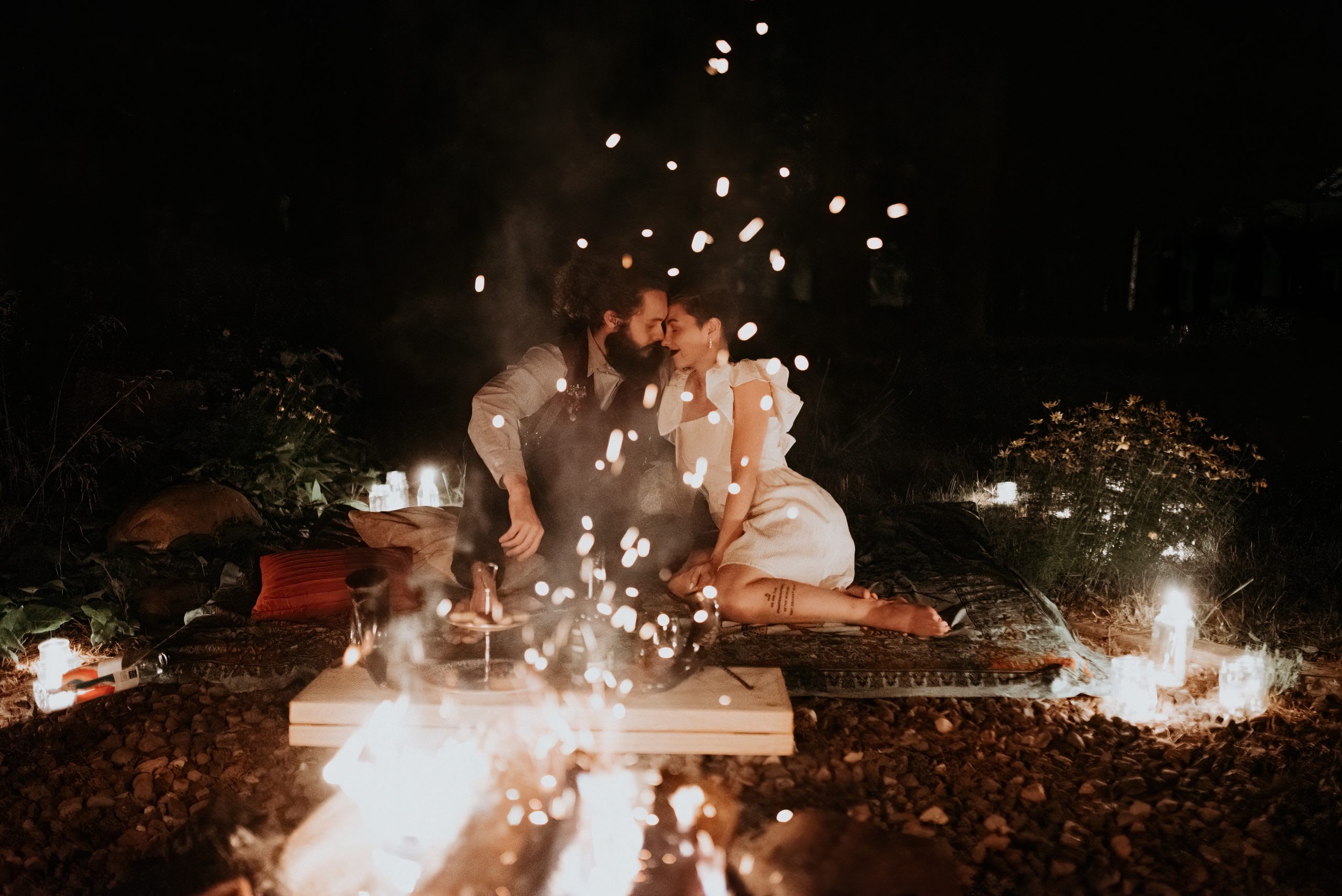 Couple sitting close together by campfire at night during Michigan elopement, sparks flying around them in romantic scene
