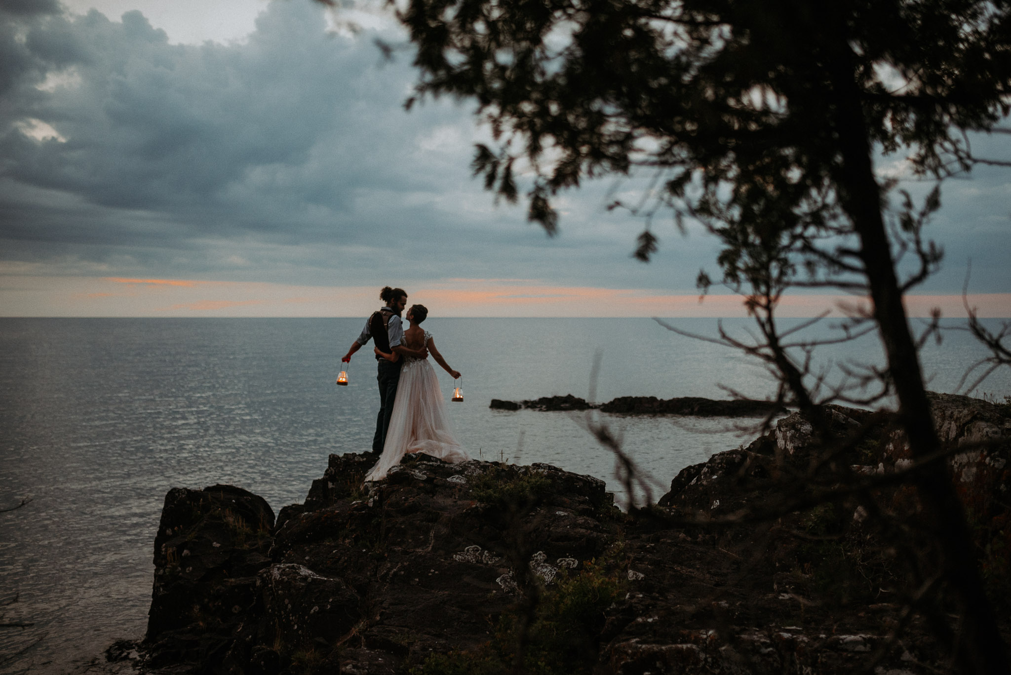 Bride and groom with lanterns standing on rocky cliff above Lake Superior under twilight sky in the Keweenaw Peninsula during Michigan elopement