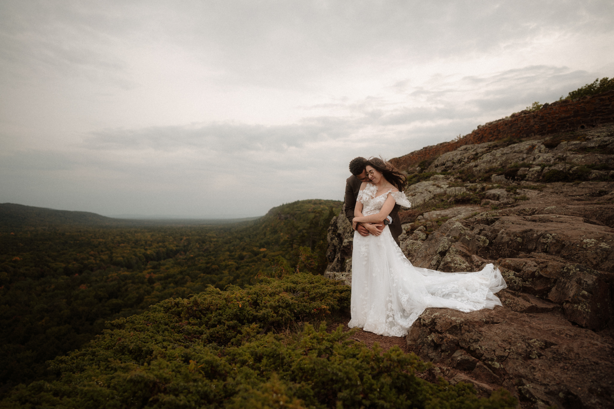 Bride and groom embracing on rocky overlook with dramatic sky at Lake of the Clouds in the Porcupine Mountains